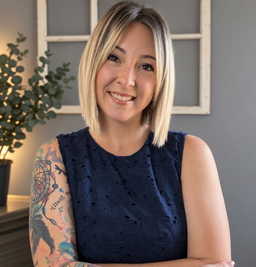 A woman with shoulder-length blonde hair smiles warmly while wearing a navy blue sleeveless top. She has colorful tattoos on her arms and stands in front of a neutral-colored wall with a plant.